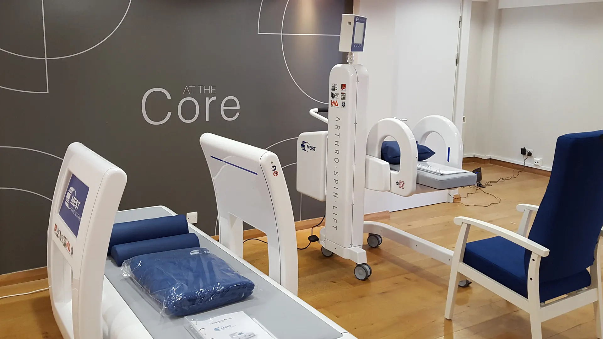 Innenaufnahme vom MBST-Behandlungszentrum At The Core Ltd in Bath. In einem großem Raum mit einem grauen Designelement auf dem der Praxisname 'At the Core' steht sind die neuen Therapiegeräte ARTHRO·SPIN·LIFT und ARTHRO·SPIN·FLEX platziert.